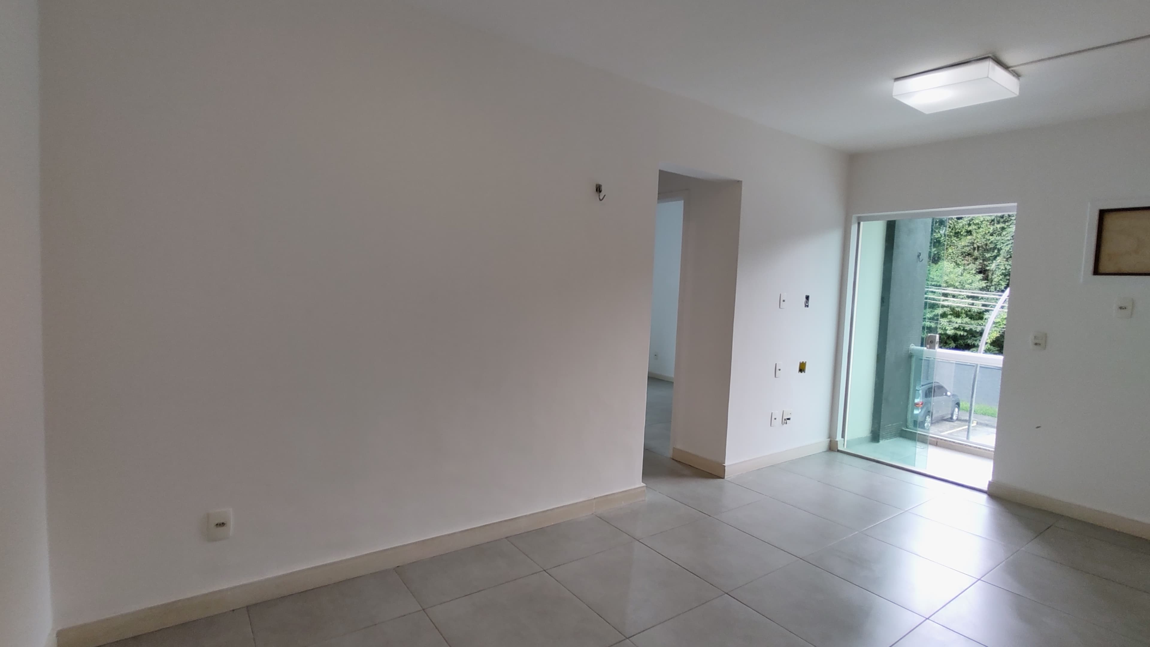 Sala vazia com piso em cerâmica clara, paredes brancas e porta de vidro com acesso à varanda.