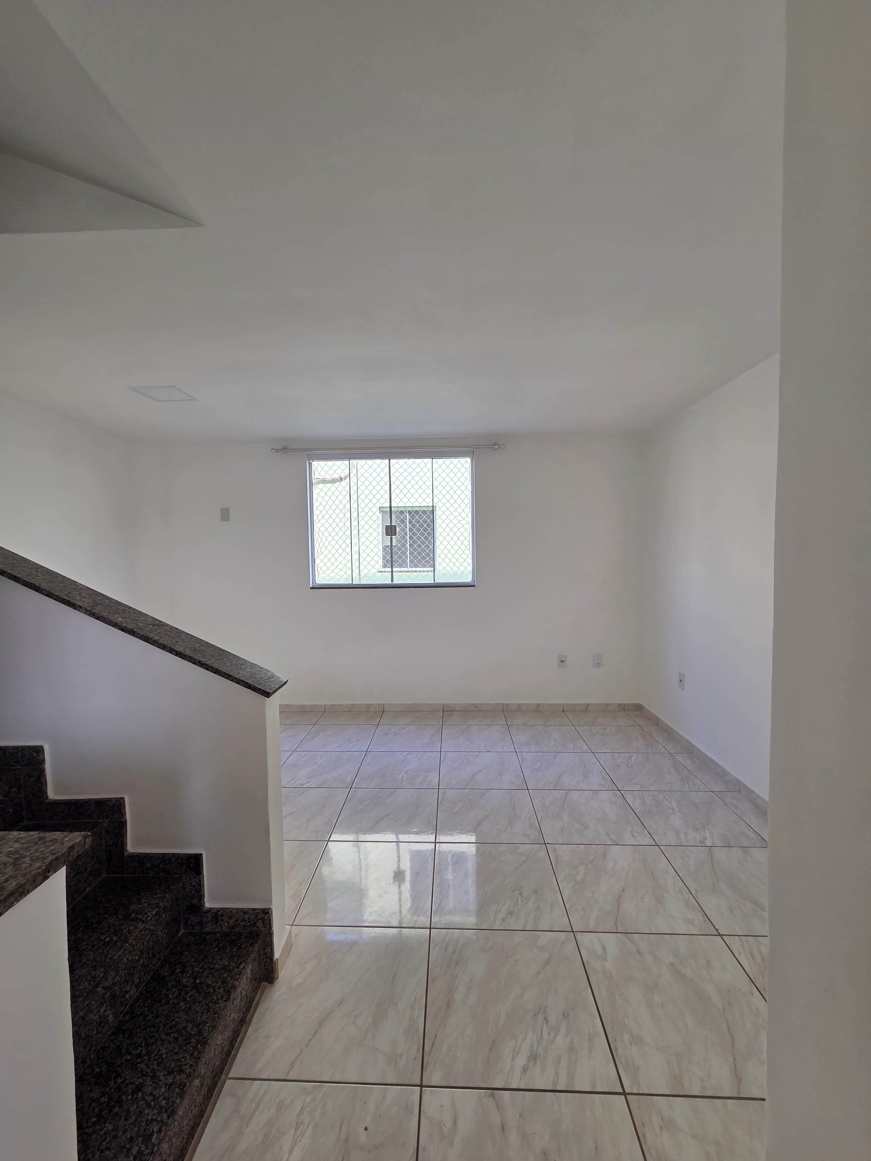 Sala ampla e vazia com paredes brancas, piso em cerâmica clara e escada com degraus em granito.