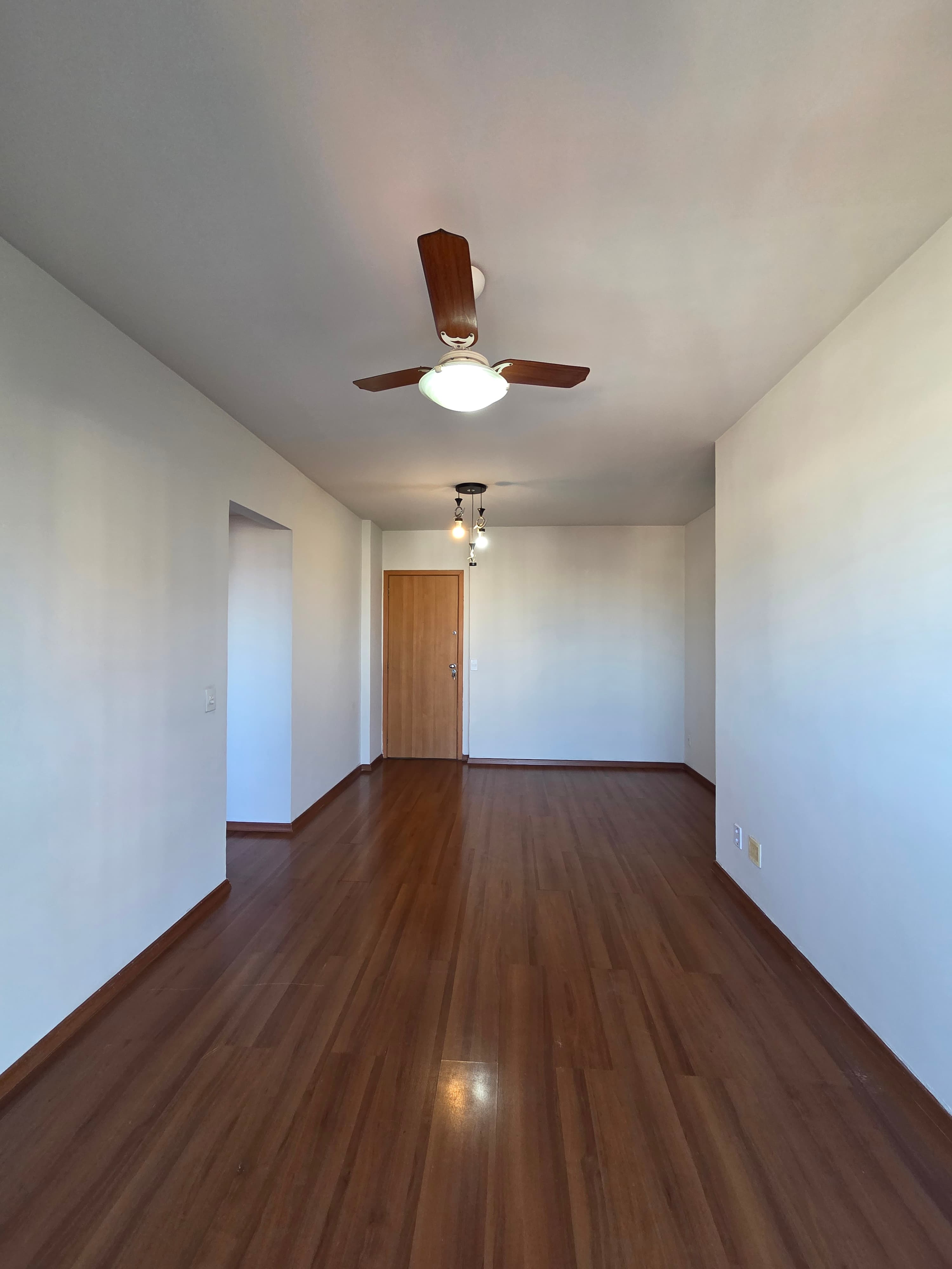 Sala ampla e vazia com piso amadeirado, paredes brancas e ventilador de teto.