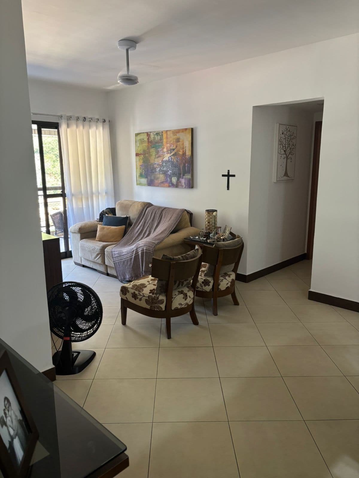 Sala de estar com piso claro em cerâmica, sofá bege com manta, duas poltronas estampadas, ventilador de teto e quadro na parede.